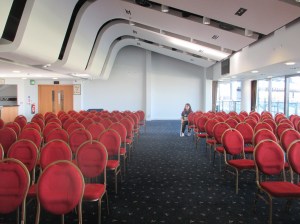 A long white room with a sloped ceiling and blue carpet. There are rows of empty red chairs.