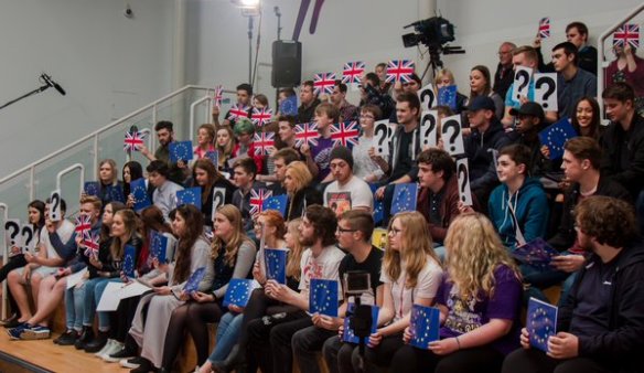 A crowd of adolescents on stepped seating. They hold up cards with Union Flags, EU Flags or Question Marks.