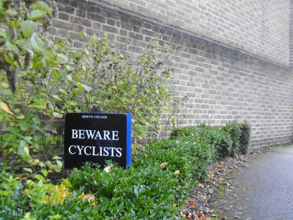 A bush-lined path with a black sign bearing the words "Selwyn College Beware Cyclists"