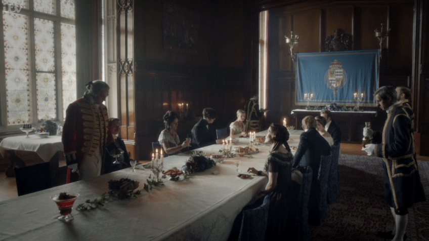 A wood-panelled dining room with a long table covered by a white sheet. Guests in luxurious costume. On the back wall is a hanging cloth printed with the arms of The Viscount Palmerston.